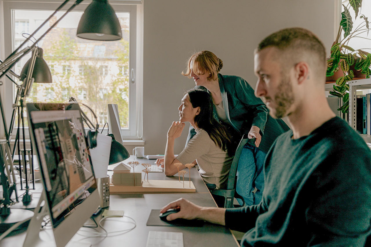 Colleagues working side by side in a bright studio, with two teammates discussing work at a screen.