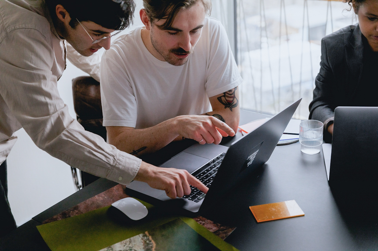 Three colleagues gathered around a table, discussing work on a shared laptop.